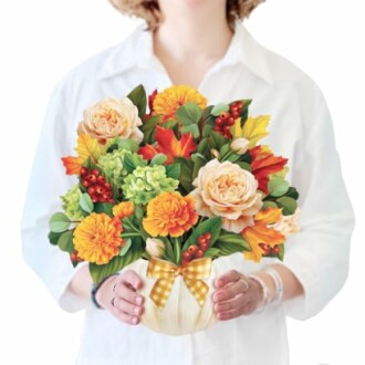 Person holding autumn-themed floral bouquet with orange and yellow flowers in a white vase.