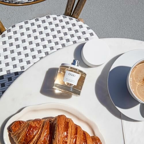 Perfume bottle, croissant, and coffee on a table.