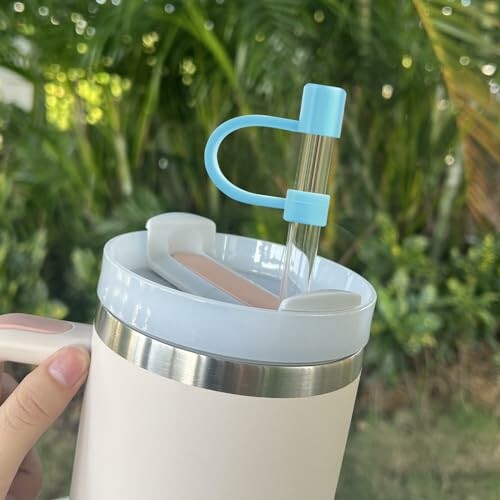 Hand holding an insulated cup with straw against a leafy background.