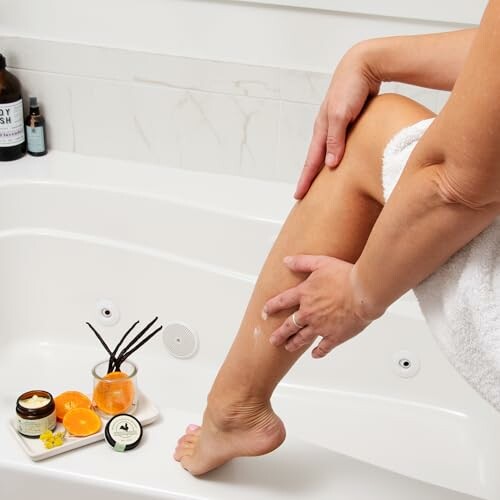 Person moisturizing leg near bathtub with skincare products.