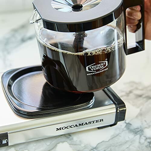 Hand holding a Moccamaster coffee pot on a warming plate.