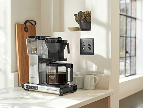 Modern coffee maker on kitchen counter with mugs and cutting board.