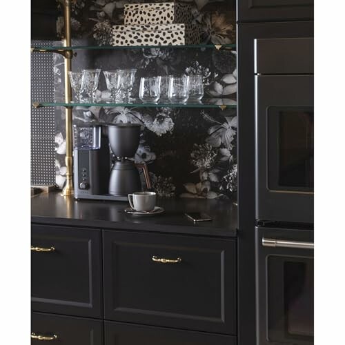 Black kitchen counter with coffee maker, cups, and patterned wallpaper.