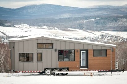 Modern tiny house on a trailer in a snowy landscape with mountains.