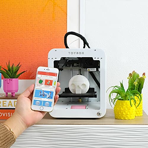 Person using smartphone to control a Toybox 3D printer on a shelf with plants.