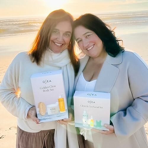 Two women holding OSEA skincare product sets on a beach at sunset